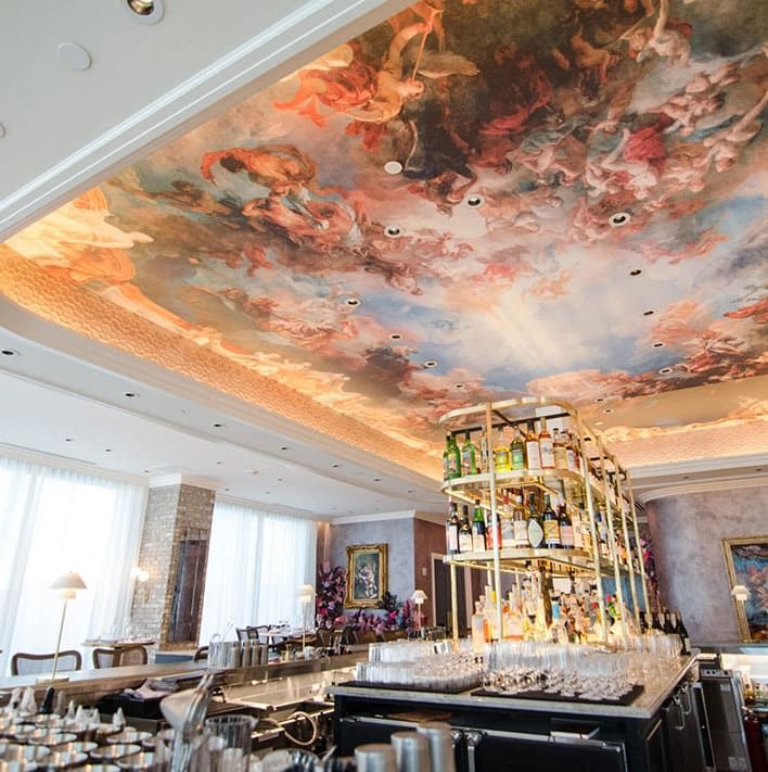 Elegant bar at Coquette Mediterranean Cuisine with a large central island, glassware, and bottles; ornate ceiling features a colorful classical-style mural with figures and clouds. Natural light filters through large windows.