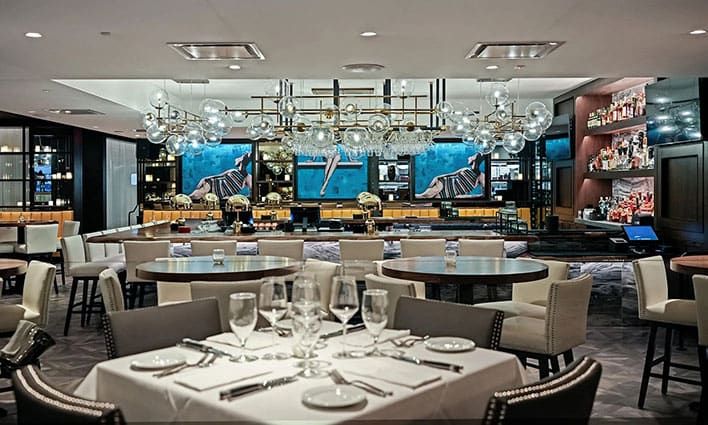 Steak 48 Del Mar, a modern restaurant interior with elegantly set tables, upholstered chairs, glassware, chandeliers, and a bar area in the background.